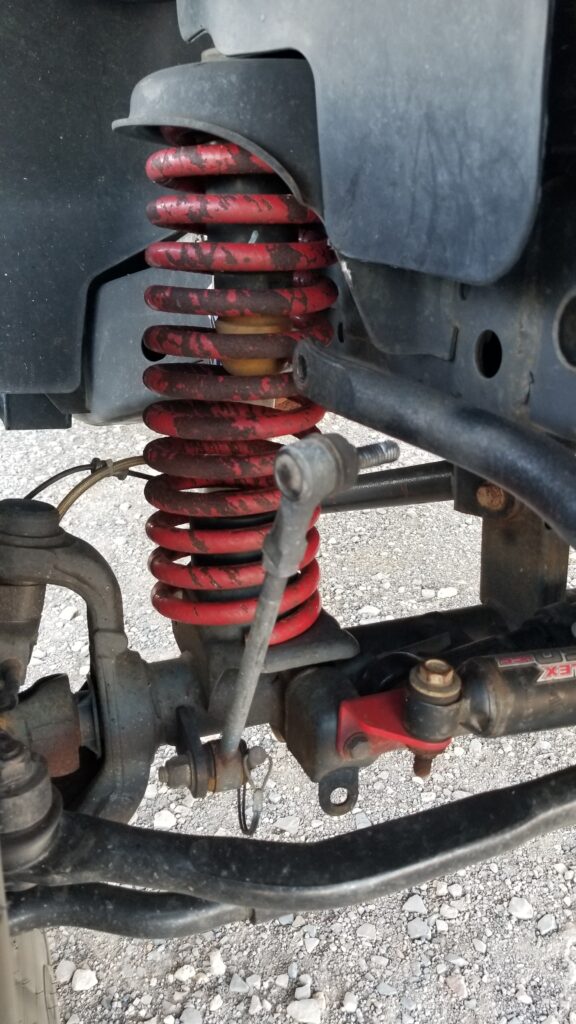 Close-up of a Jeep Wrangler's front suspension showing a red coil spring, sway bar, and a disconnected sway bar end link with a bolt missing. The suspension is under no load, and the loose end link is hanging, illustrating a common off-road issue when hardware backs out or loosens.