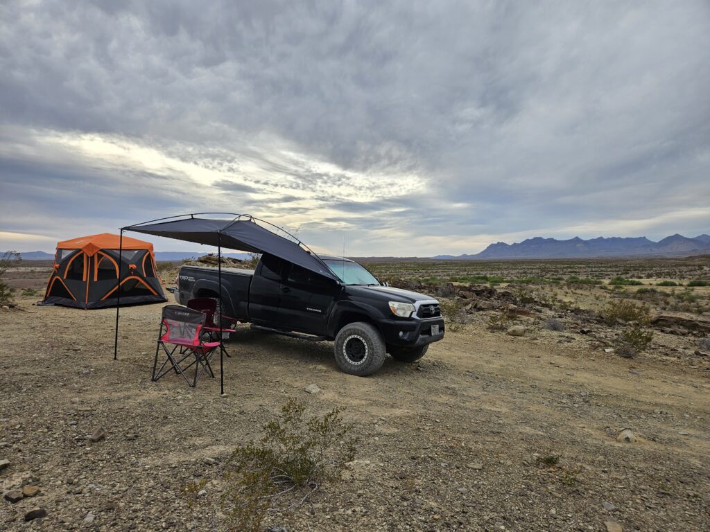 Backcountry overlanding campsite at Big Bend National Park featuring a black Toyota Tacoma with awning setup, orange tent, and desert mountain landscape under overcast skies.
