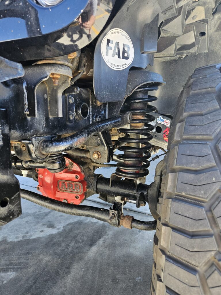 Close-up of a Jeep Wrangler's front suspension showing a red ARB differential cover, coil spring, and sway bar. The driver-side sway bar end link is missing, leaving the sway bar unattached on one side. The image highlights a common off-road maintenance issue.