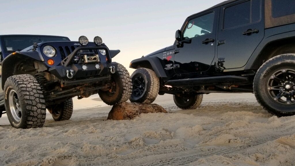 Thanks for the clarification—here’s a corrected version: **Alt text:** Two Jeep Wranglers flex on a beach, each with one front tire on a buried log. The Jeeps face each other in a mirrored pose, showing off front suspension articulation and off-road capability in a sandy environment.
