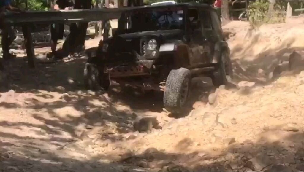 A Jeep Wrangler navigating a deeply rutted, rocky trail section under tree cover. The front suspension is visibly compressed on one side and drooping on the other, highlighting articulation in rough terrain. Several people observe from behind a guardrail in the background.