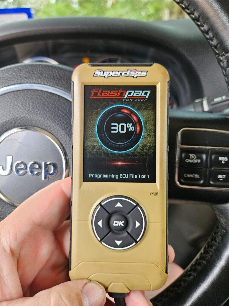 Hand holding a Superchips Flashpaq tuner while programming the ECU on a Jeep Wrangler, showing 30% progress on the screen.